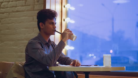 young man in gray shirt sipping drink from disposable cup with straw while sitting beside large window glowing with soft blue outdoor light, relaxed mood in cozy cafe with rustic interior and warm ambiance