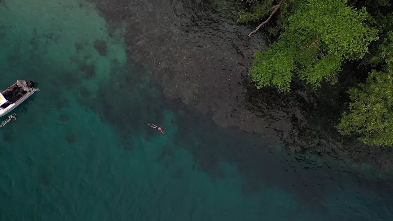 한 젊은 백인 남자가 그의 작은 흰색 보트 옆에 있는 산호초 위의 수면에서 지느러미를 가지고 수영하고 있는 공중 샷