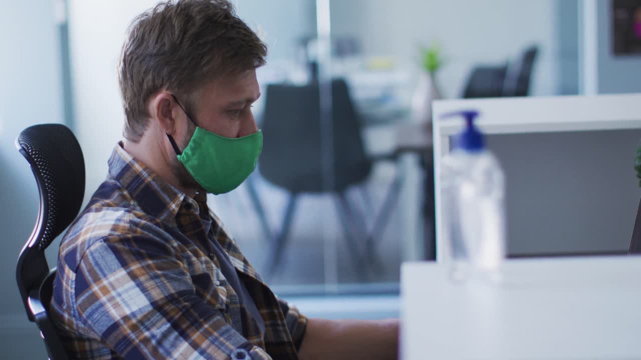 hombre caucásico con máscara facial en la oficina, usando una computadora portátil