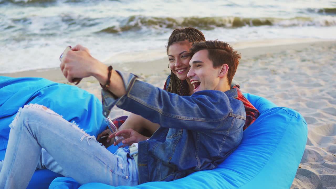 una pareja divirtiéndose y tomando selfies acostados en sillas cómodas con una manta en la playa. filmado en 4k.