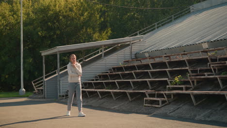 young lady in sporty attire, walks thoughtfully near a stadium on a sunny day, surrounded by lush greenery and electric poles, she appears pensive, with the empty stadium seating environment