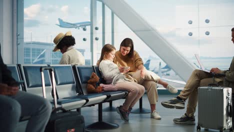 terminal de avión ocupado del aeropuerto: madre hermosa y hija pequeña encantadora esperan su vuelo de vacaciones, usan teléfonos inteligentes móviles para divertirse. grupo diverso de personas en la sala de embarque del centro de la aerolínea