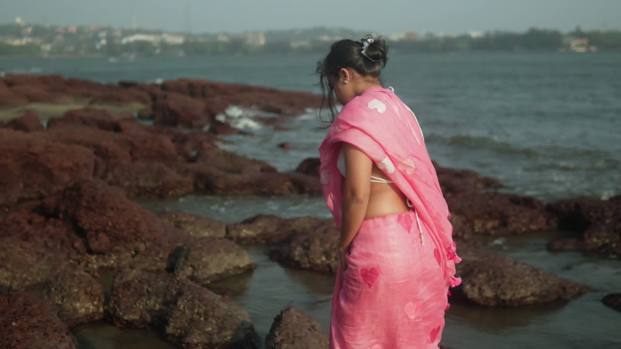 Donna in sari rosa che cammina su una costa rocciosa, con le onde che si infrangono, atmosfera serena, vista laterale.