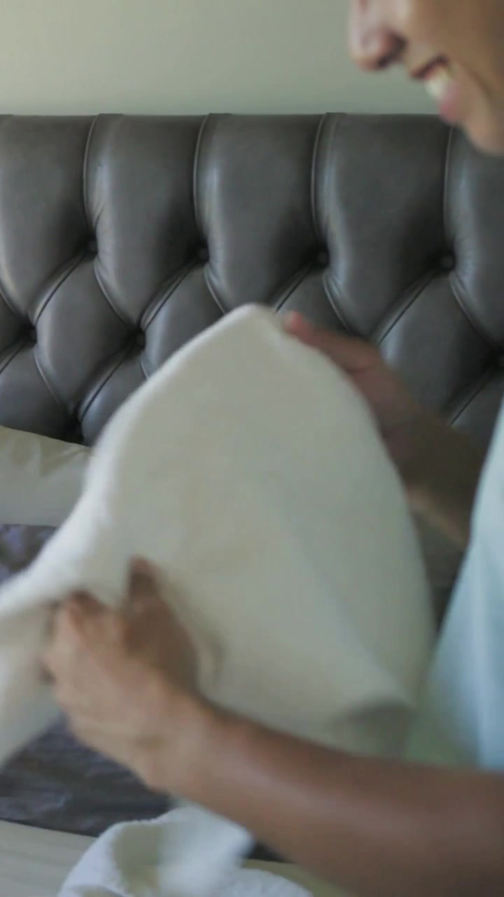 Video verticale: Uomo in maglietta che prende un lenzuolo, lo dispiega e liscia la biancheria vicino a una testiera capitonné per rifare il letto.