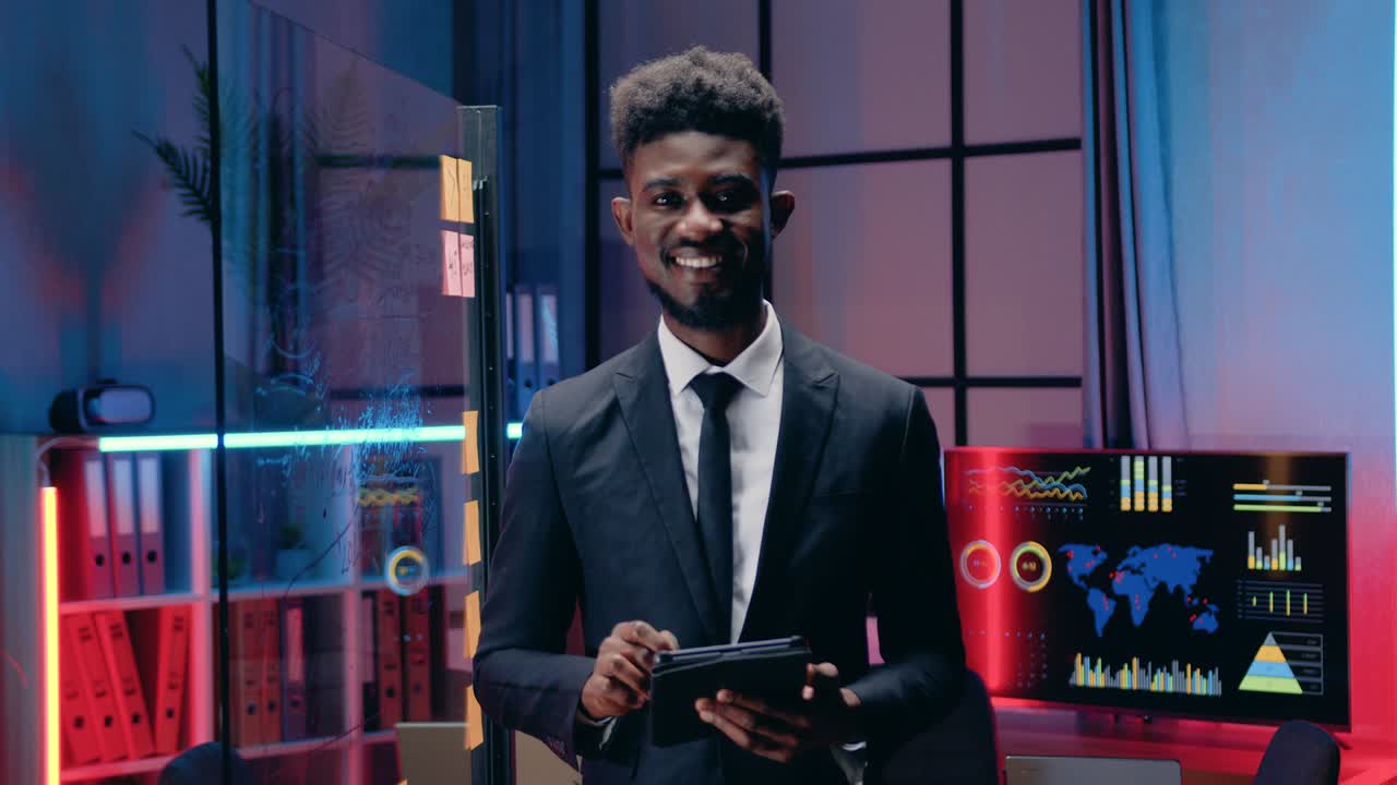 agradable positivo seguro de sí mismo decidido hombre de negocios africano barbudo trabajando en una tableta en la oficina de la noche y luego mirando a la cámara con una sonrisa sincera, vista frontal