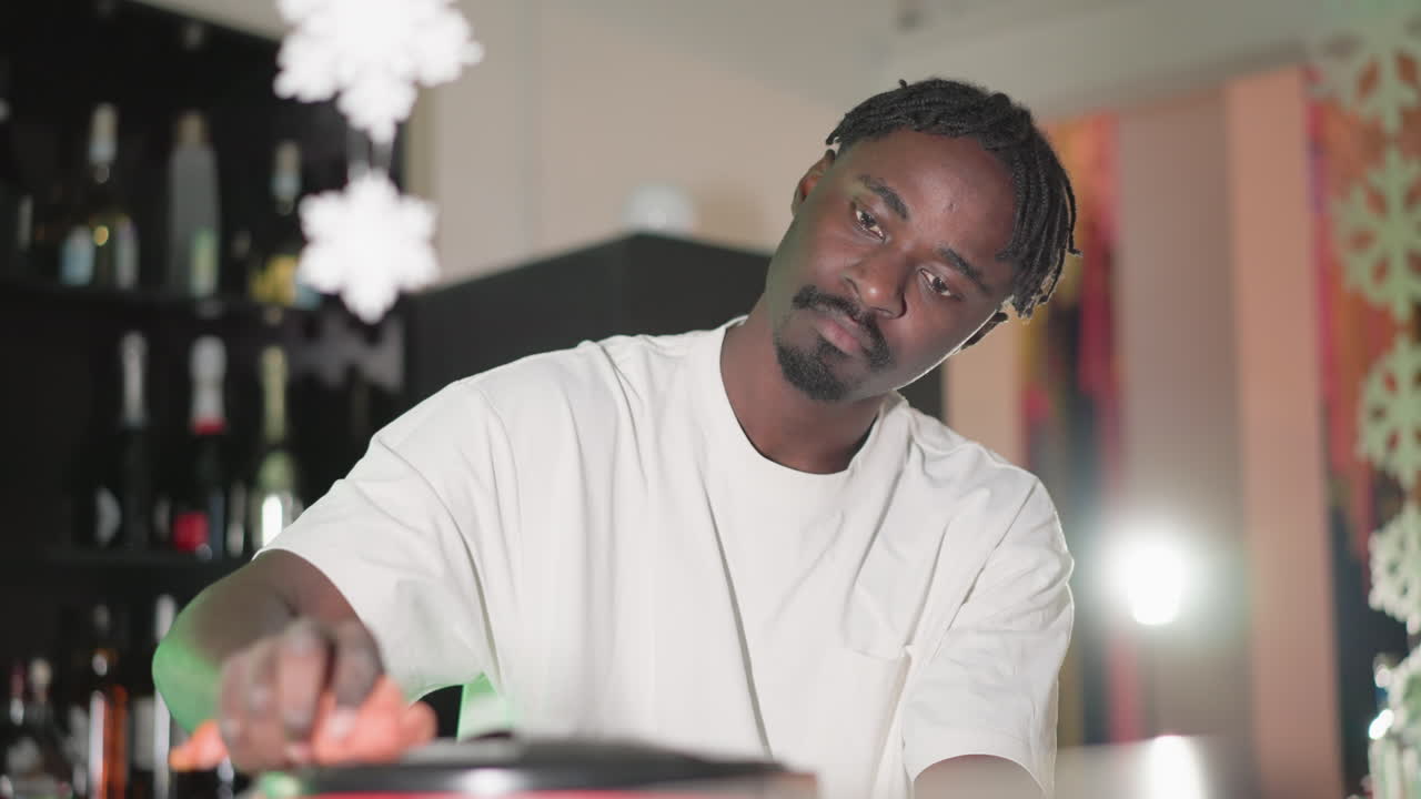 Bartender focused on wiping surface of trash bin behind bar counter. Wearing a white t-shirt, the bartender s hand reaches for the lid while green neon lighting from Heineken sign adds to the ambiance