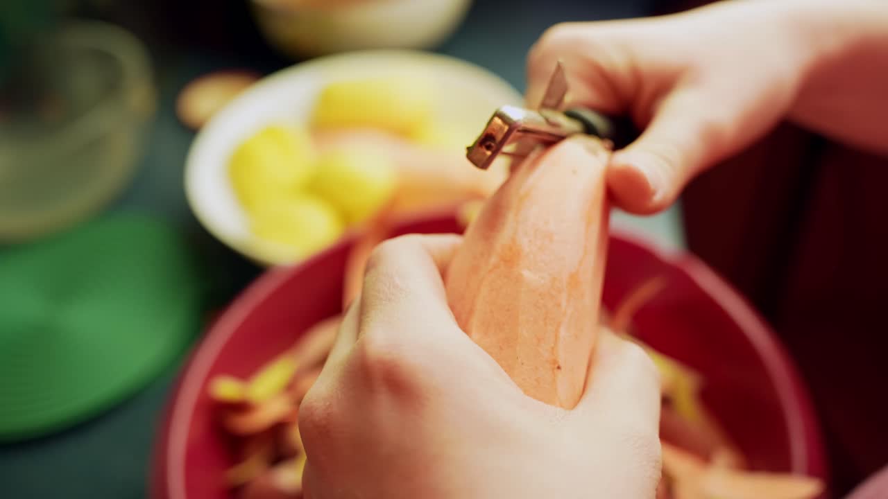 Primo piano di mani che usano un pelapatate per sbucciare una patata dolce sopra una ciotola su un bancone della cucina, scena di calda preparazione di cibo casalingo.