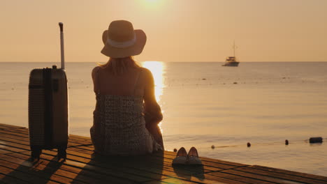 a woman looks at the ship in the distance sitting on the pier near her luggage the travel concept