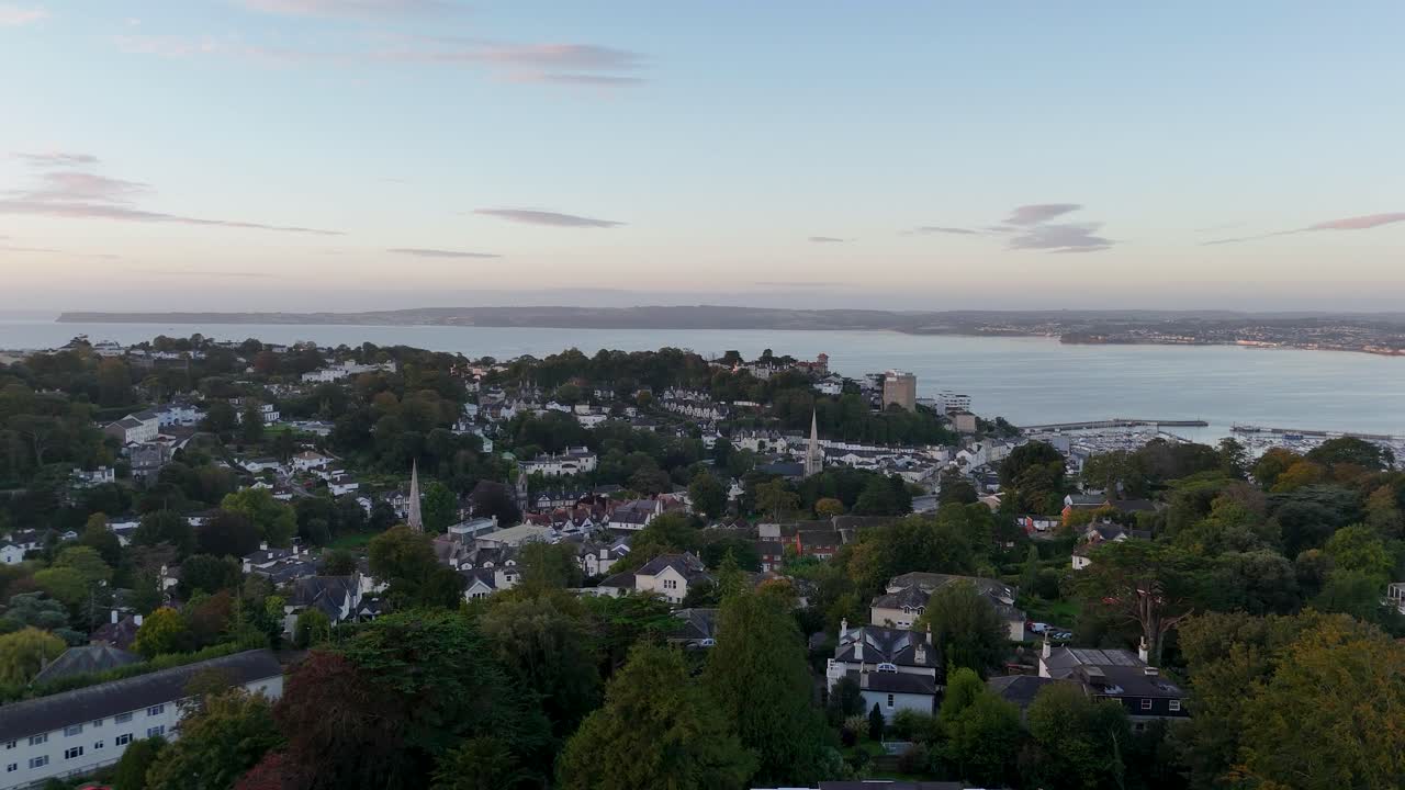 Cinematic aerial drone push in movement over residential houses and trees revealing coastal bay and waterfront in Torquay Devon England during dawn with soft evening light and tranquil atmosphere