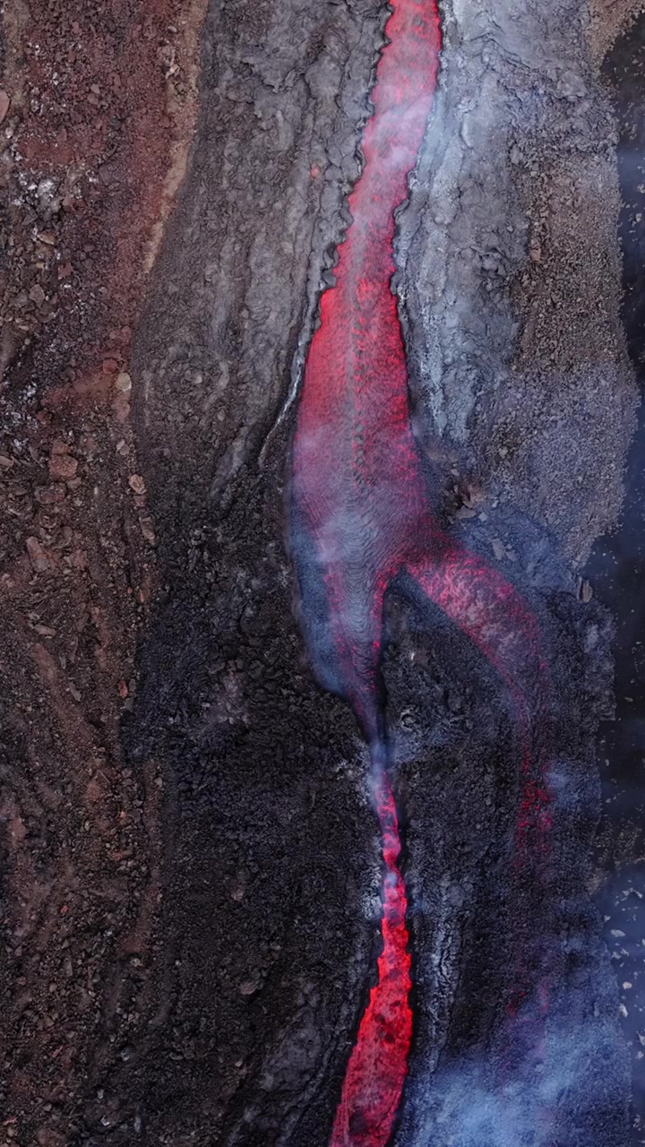 Vista aerea di un fiume di lava incandescente che scorre attraverso un terreno vulcanico scuro, con vapore che sale