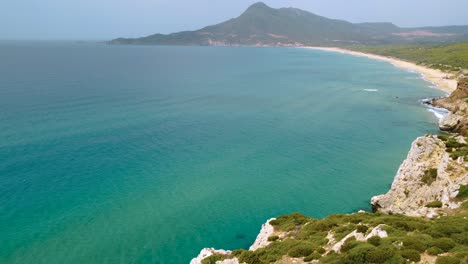 Aerial-of-an-idyllic-natural-rocky-coast-beach-sandy-bay-on-the-tourist-vacation-island-Sardinia,-Italy,-with-mountains,-clear-blue-turquoise-and-calm-water-close-to-Costa-Rei