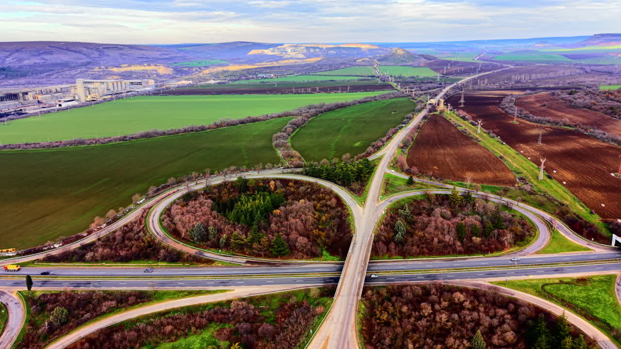 Eine komplexe Straßenkreuzung mit mehreren Auffahrten, umgeben von landwirtschaftlichen Feldern und Industriegebäuden in der Ferne. Luftaufnahme eines Autobahnkleeblatts auf dem Land.