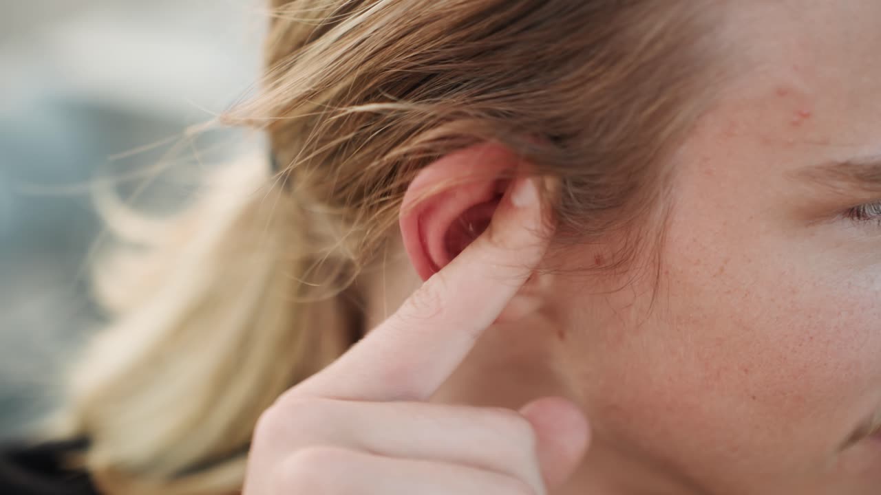 Primo piano: uomo bianco che si massaggia l'orecchio irritato, con cartilagine rossa visibile. Il polpastrello applica pressione vicino al condotto uditivo. In sottofondo, una brezza marina e acqua sfocata. L'uomo ha un'espressione di disagio, in un momento di autocura.