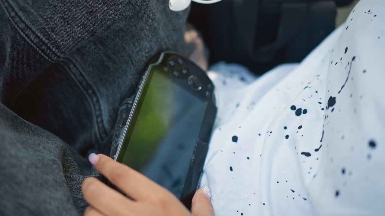 primer plano de unas manos desenredando el cable de los auriculares sobre el regazo, camisa moteada y vaqueros visibles, reloj en la muñeca, dedos manipulando el cable, reflejo de la pantalla visible, movimiento táctil cuidadoso, ritual de audio íntimo
