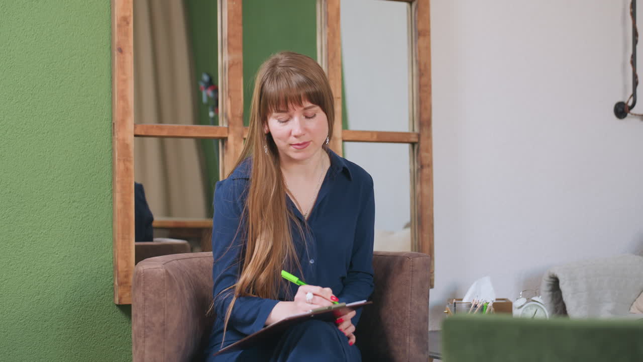 Female artist seated on sofa writing with green pen on notepad, lifting face with gentle smile in cozy indoor room with wooden mirror, pen holder, tissue box, and small clock on table nearby