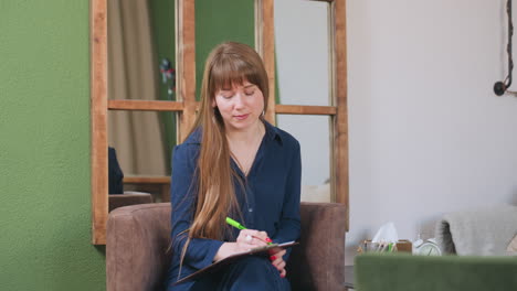 female artist seated on sofa writing with green pen on notepad, lifting face with gentle smile in cozy indoor room with wooden mirror, pen holder, tissue box, and small clock on table nearby