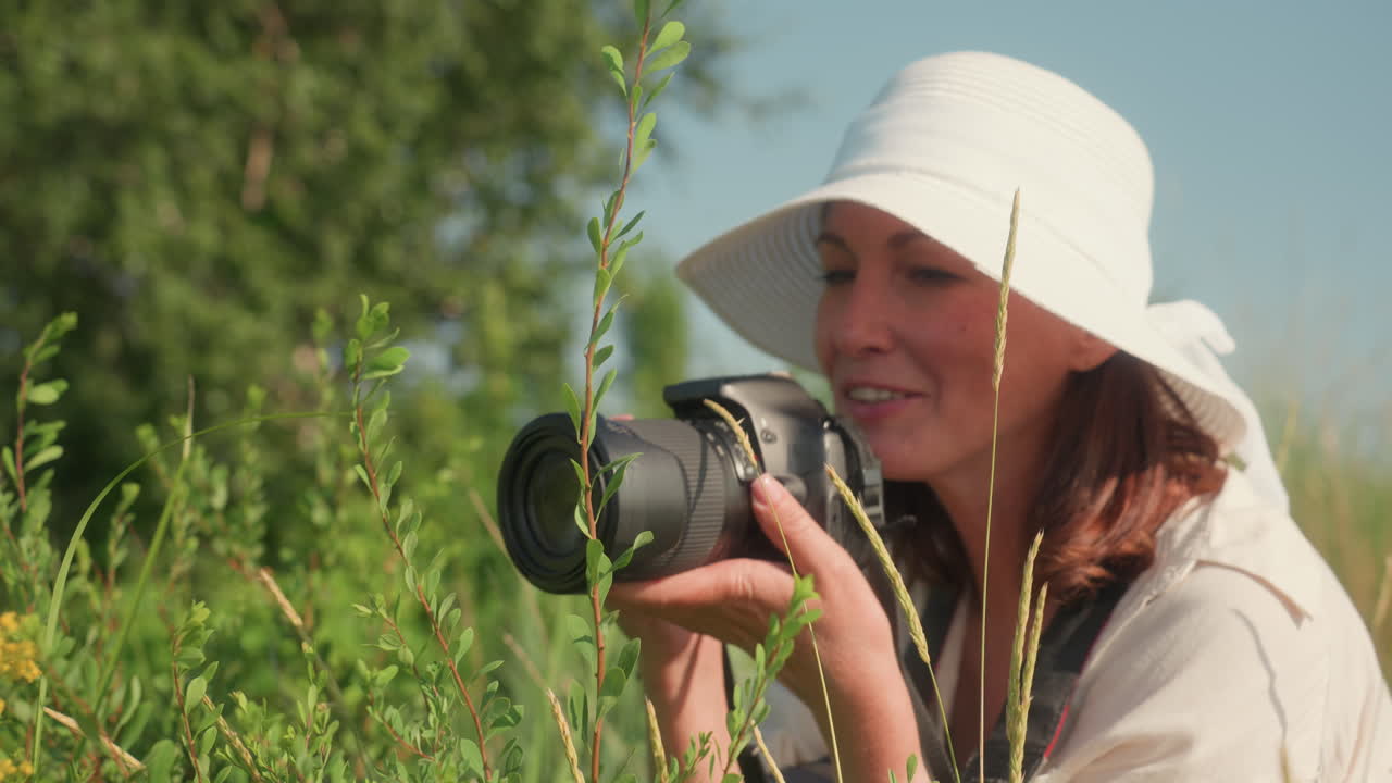 Smiling woman in white sunhat crouches in tall grass with camera in hand preparing to photograph wildflowers under bright daylight in lush green meadow