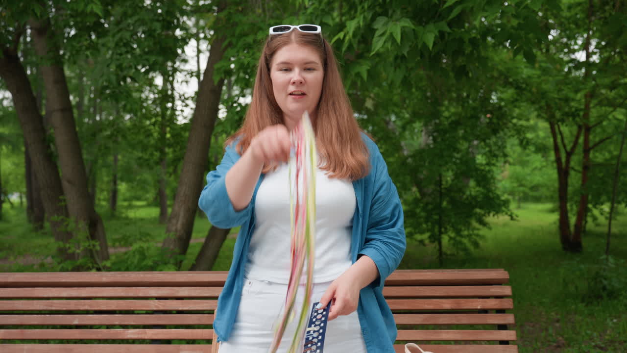 흰색 옷과 파란색 셔츠를 입고 밝은 표정으로 공원에서 알록달록한 자수 실을 장난스럽게 던지며 미소짓고 있는 젊은 여성, 명랑한 창의성을 보여줘요.