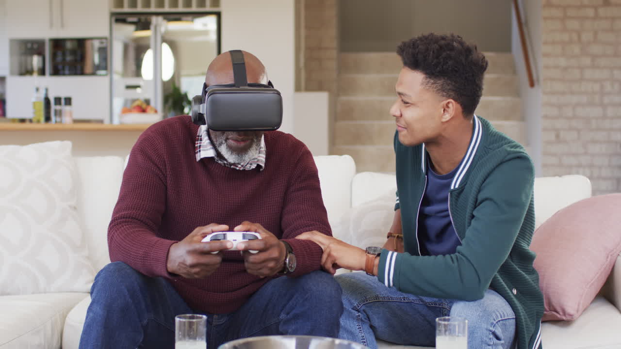 Happy african american father using vr headset and gamepad with adult son at home, slow motion
