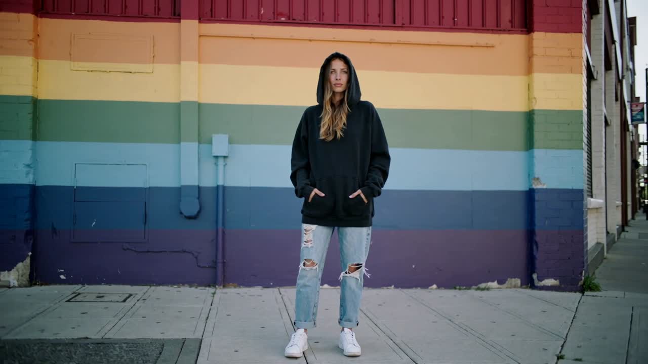 Camera starting, woman posing and shifting posture on city sidewalk by rainbow wall, hoodie up