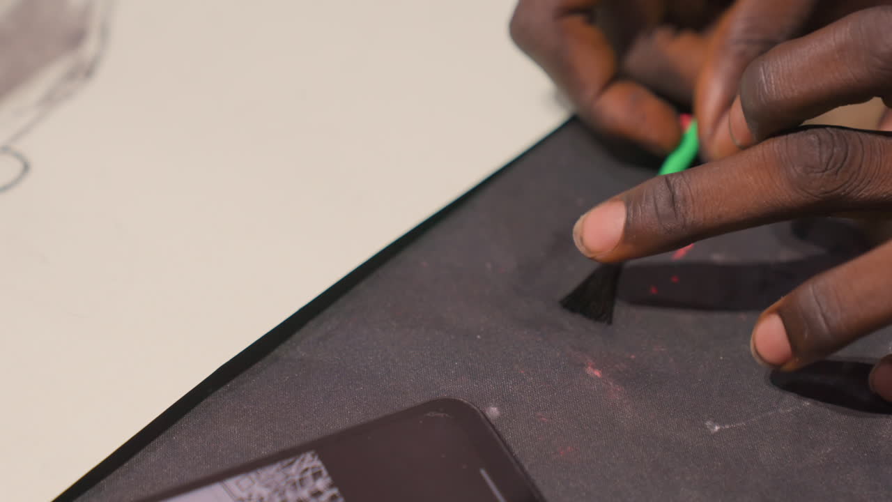 Closeup hands etching black painting mat, artist using green tool to scrape pigment from textured surface next to smartphone, red paint specks, precise motion, focused tactile studio atmosphere