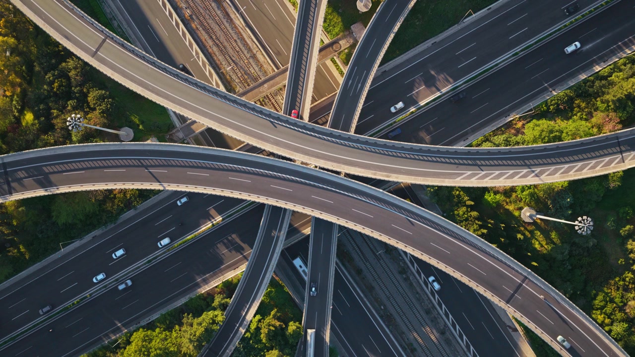 Aerial top-down view of a massive highway intersection with multiple overpasses and bridges, showing cars and trucks driving along the intricate road network amidst green trees at sunset