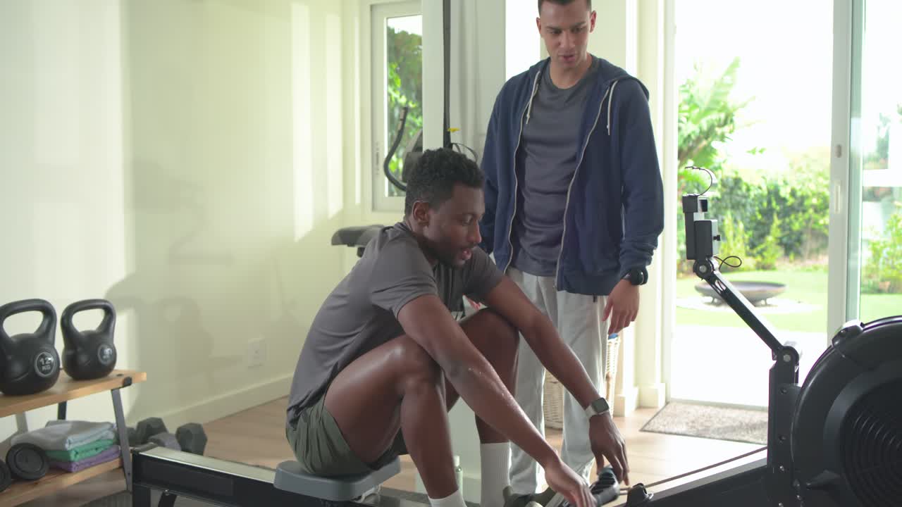 Coach entering home gym checking smartwatch coaching African American man rowing and offering water
