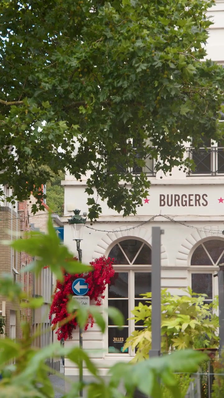 La cámara se desliza entre ramas frondosas, revelando la encantadora fachada de un restaurante de hamburguesas en un callejón histórico de Brujas.