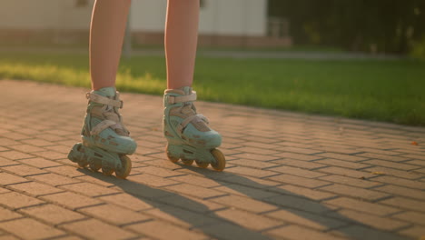 leg view of lady roller skating slowly on interlocked path, wearing teal rollerblades, casting shadow on ground under warm sunlight, blurred background featuring lush greenery, building and trees