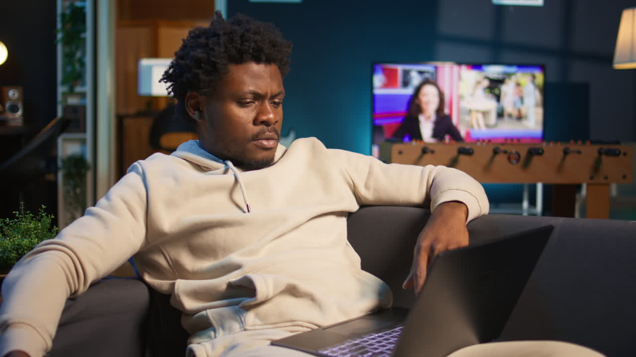 Adult man typing on a computer at his stylish illuminated apartment, engaging in online work, multitasking efficiently and embracing the flexibility of remote work in a cozy home setting. Camera B.