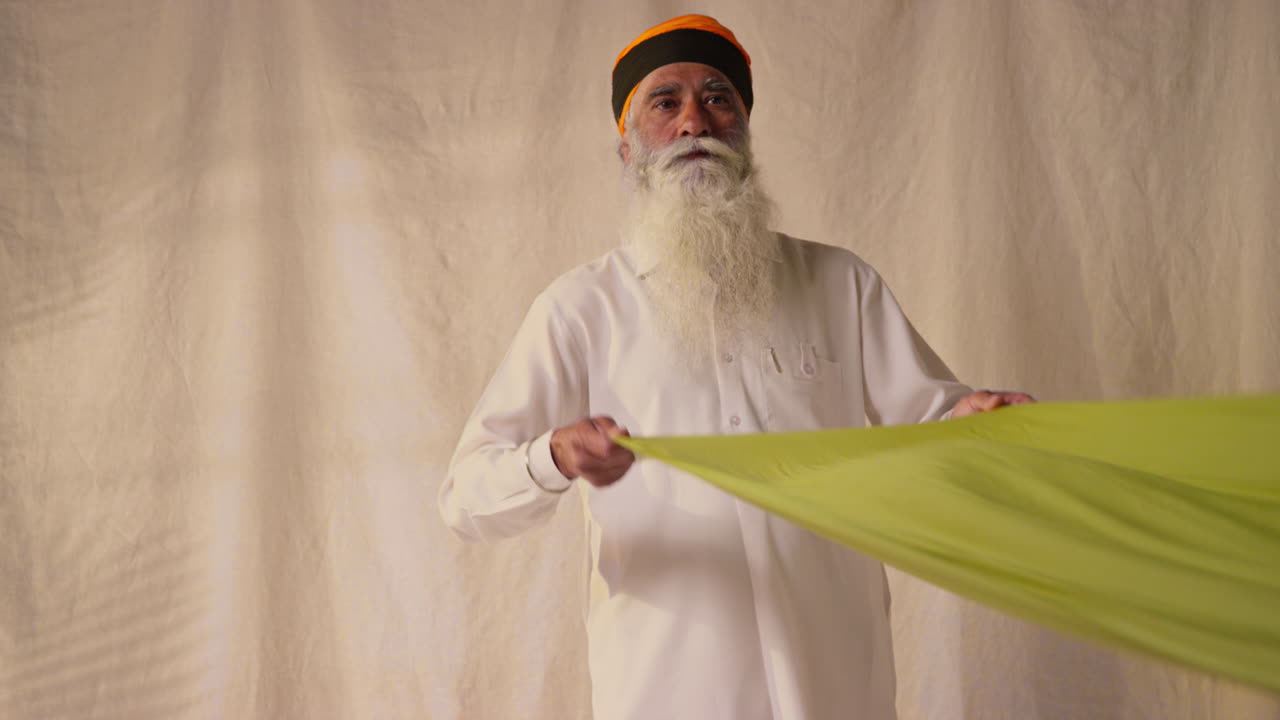 Studio Shot Of Senior Sikh Man Folding Fabric For Turban Against Plain Background As Sequence Part 2 Of 2