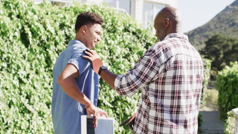 african american father talking to adult son holding solar panel in sunny garden, slow motion