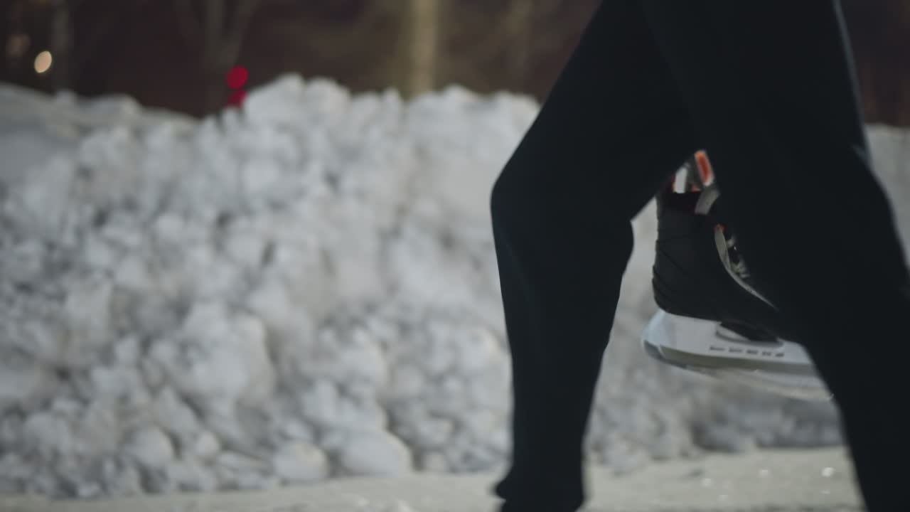 Side view of skater walking outdoors holding black skating boot near large icy snow pile as bokeh lights filter through bare tree in background