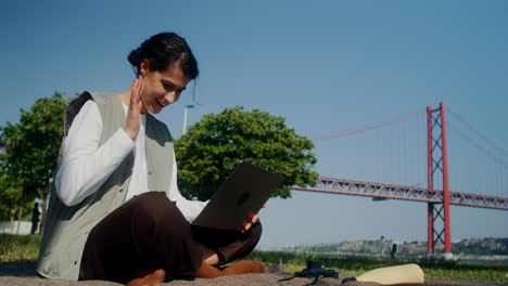 woman working on a laptop in a park near a bridge