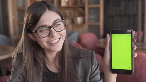 Adorable-Joven-Mujer-De-Negocios-Con-Anteojos-Está-Sentada-En-Un-Café,-Sosteniendo-Un-Teléfono,-Sonriendo-Y-Demostrando-Una-Pantalla-Verde-Del-Teléfono