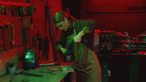 caucasian young woman welder in apron working with electrodrill and drilling in workshop