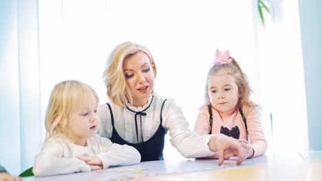 cute preschoolers and their teacher at the table. blonde teacher controls the work of kids and helps them to study. woman plays with little children together indoors.