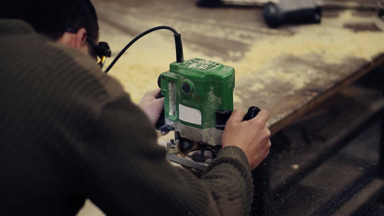 나무 작업장에서 수동  ⁇ 는 기계로 일하는 보호 안경의 마스터. 큰 나무 판을  ⁇ 습니다. 먼지와  ⁇ 이 바닥에  ⁇ 어지고 있습니다. 느린 움직임. 어깨에서 촬영