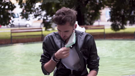 a young man with a fashionable hairstyle wearing a black leatherette jacket, white t-shirt, sitting on a fountain in a park, removing a green protective covid-19 mask from his face, close up 4k shot