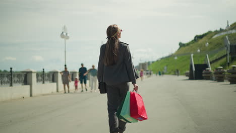 dama con gafas de sol lleva bolsas de compras rojas, menta y verdes, balanceándolas suavemente mientras mira hacia la colina, con personas borrosas que se acercan desde la dirección opuesta en el fondo