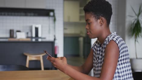 African-american-businesswoman-wearing-face-mask-sitting-in-workplace-kitchen-using-smartphone