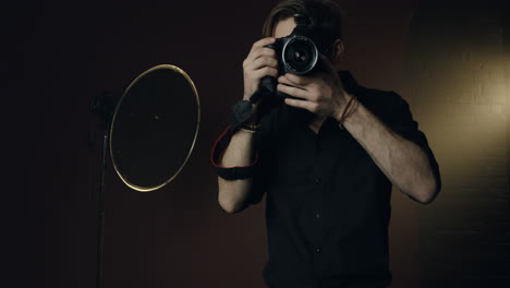 vista frontal de un joven fotógrafo caucásico tomando una foto con una cámara y sonriendo en el fondo oscuro del estudio
