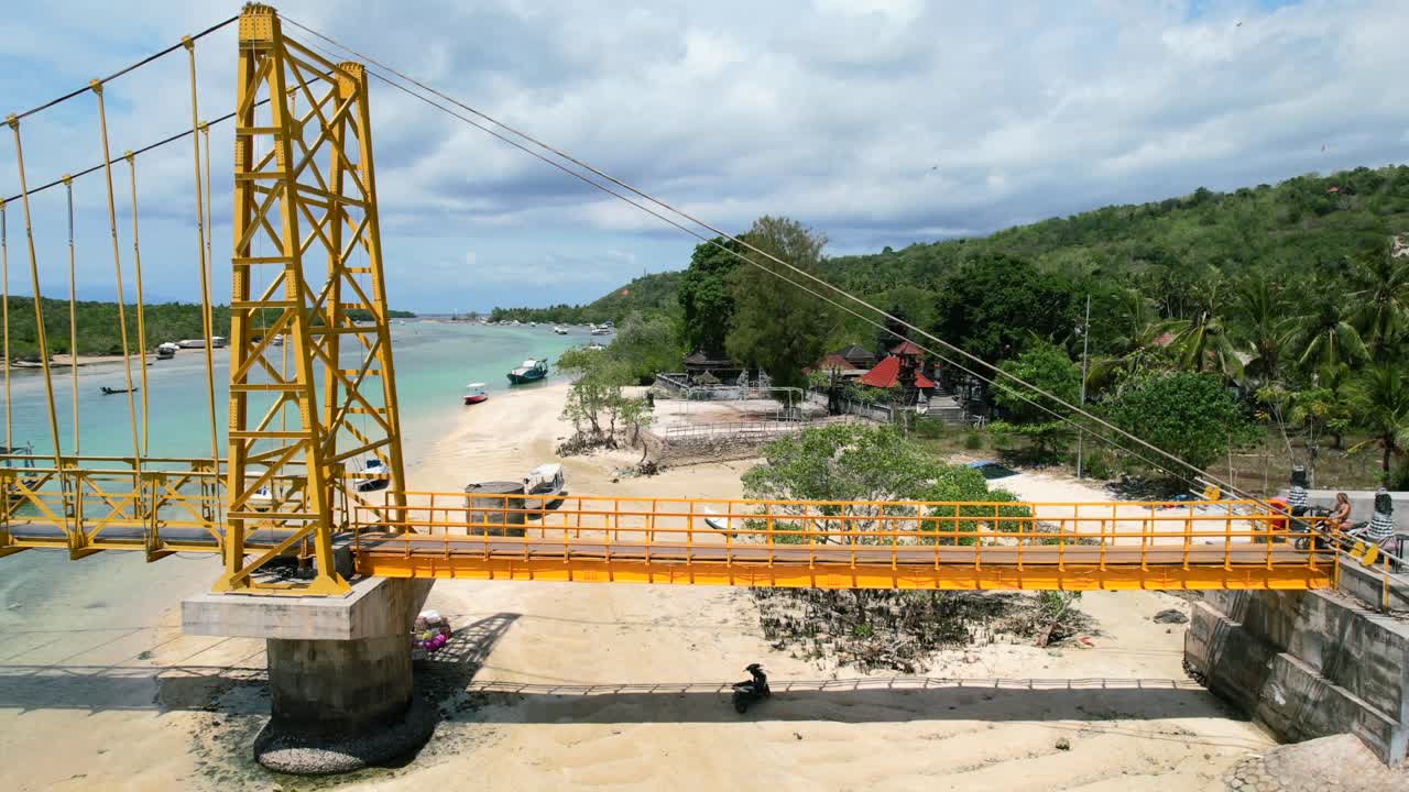 공중 삼각대는 화창한 여름날 인도네시아 발리 누사 세닝간에서 노란색 현수교를 가로질러 운전하는 스쿠터에서 축소됩니다.