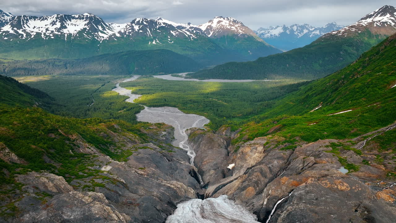 Glacier descends into the gulch in the rocks and transferring into a small river flowing in the valley. Strikingly beautiful mountains with snow on the slopes at backdrop. Nature of Alaska, USA. Aerial view