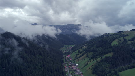 Vista-Aérea-A-Lo-Largo-De-Un-Valle-Con-Un-Pueblo-Turístico-De-Montaña,-Altas-Montañas-Con-Bosques-De-Abetos-Y-Nubes-Muy-Bajas