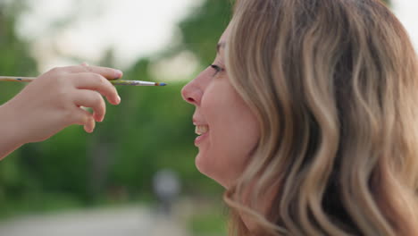 side close up young woman smiling turning face away from brush pointed at cheek during outdoor painting session on wooden dock with water reflection and trees background in soft evening light