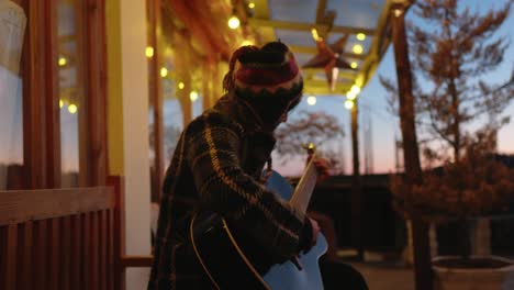 side view of a woman playing guitar on a porch at twilight with string lights