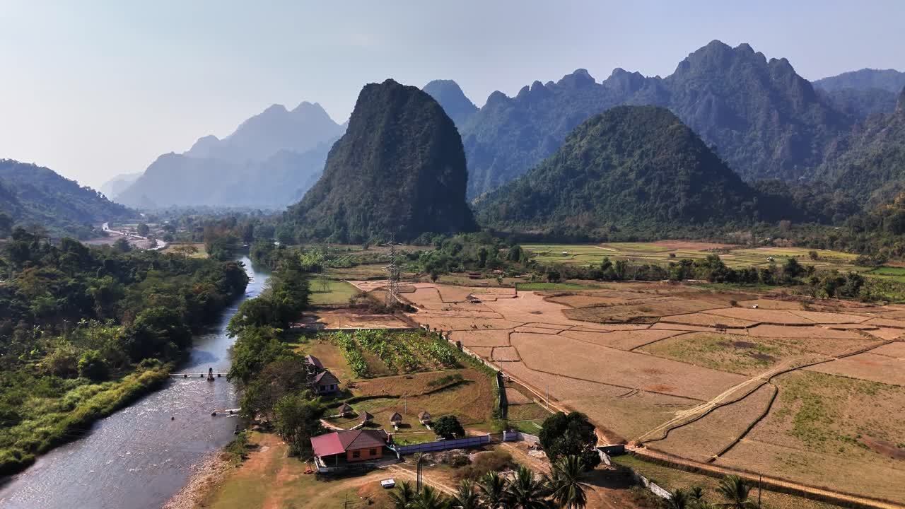 Drone captures the dramatic limestone karst formations of Pha Tang, with the Nam Song River meandering through golden rice fields and village houses near Vang Vieng in northern Laos