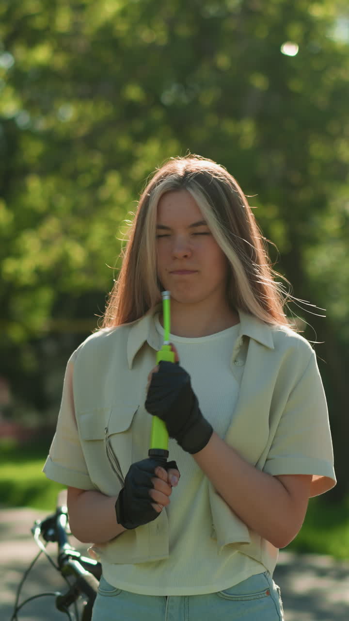 joven con cabello rubio se encuentra a la luz del sol, concentrándose mientras sostiene la bomba de aire verde cerca de la cara, sintiendo la brisa y el aleteo del cabello, el fondo muestra una escena tranquila del parque con árboles vibrantes