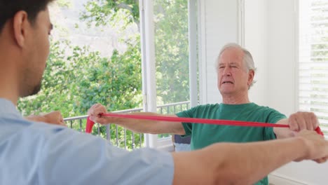 Un-Hombre-Mayor-Caucásico-Sosteniendo-Una-Banda-De-Resistencia-Haciendo-Ejercicio-Con-Un-Fisioterapeuta-Masculino-En-Un-Hogar-De-Ancianos